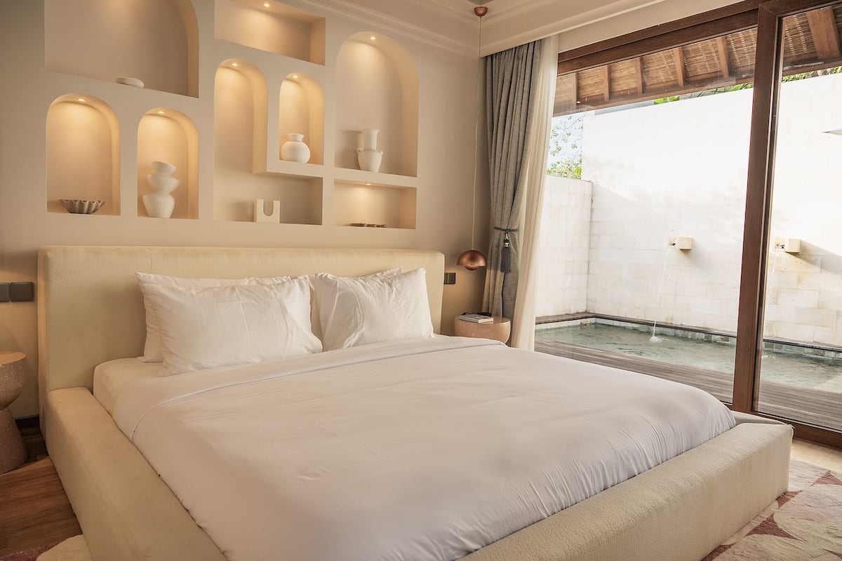 A large bed with white bedding, a bedside table with a book, and a small shelf with decorative items.