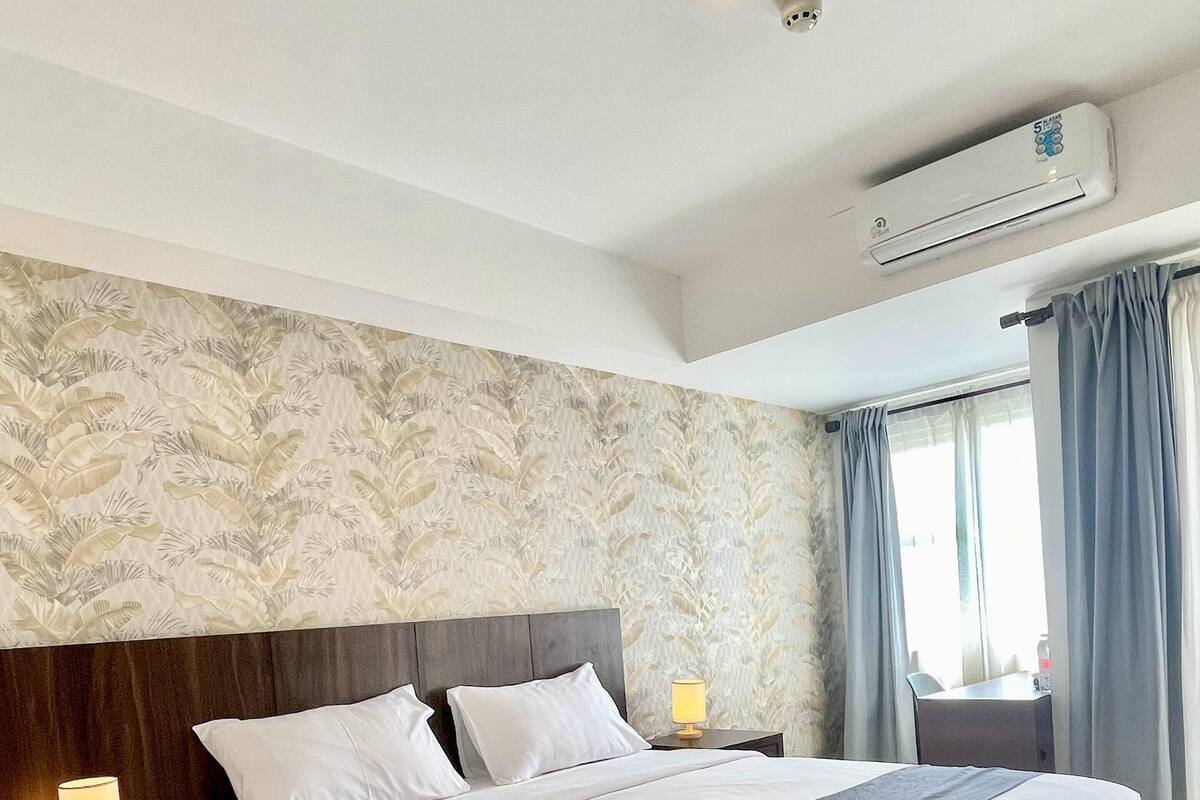 A hotel room with a large bed, a nightstand with a lamp, a wall-mounted air conditioner, and a window with curtains.