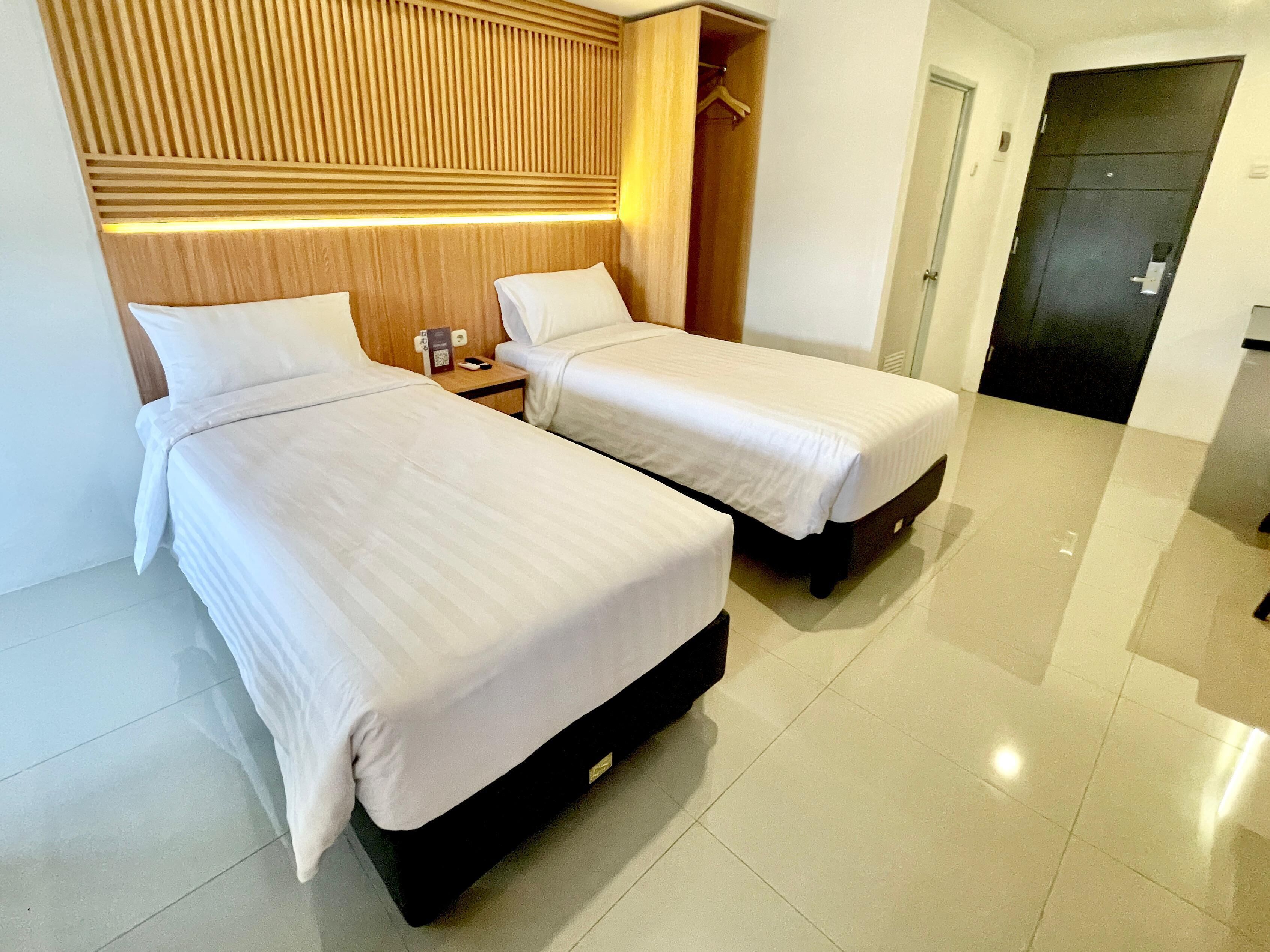 Habitación de hotel con dos camas individuales, cabecera de madera, mesita de noche con teléfono y cargador, y una puerta a la derecha.