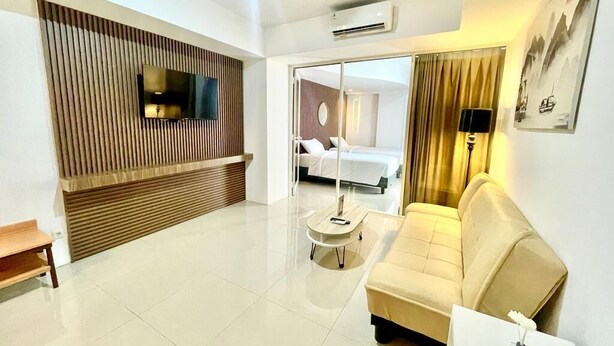 A modern hotel room with a flat-screen TV mounted on a wooden panel wall, a sofa, a small round table, and a bed visible through a glass door.