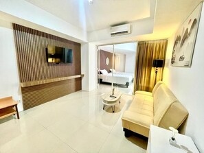 A modern hotel room with a flat-screen TV mounted on a wooden panel wall, a sofa, a small round table, and a bed visible through a glass door. - Nemuru Hotel Ciputat (South Tangerang)