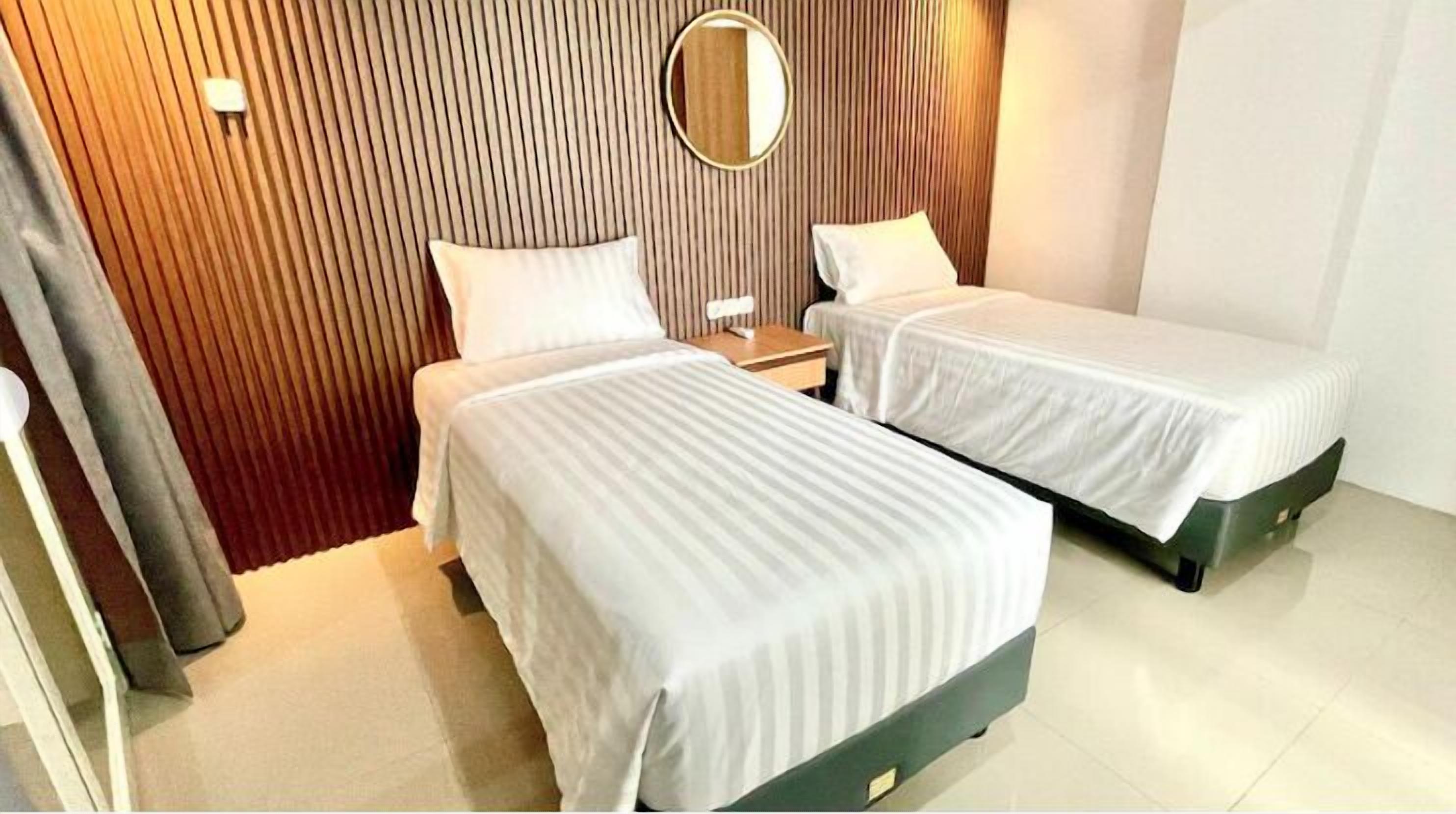 A hotel room with two single beds, a wooden slatted headboard, a round mirror, and a small bedside table.