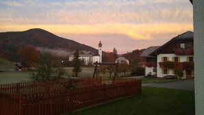 A rural setting with a white church and residential buildings, surrounded by a wooden fence and a grassy field. - Ferienwohnung Zuhaus, 76m², 2 Schlafzimmer, Balkon, TV, 4 Sterne, 4 Pers (Anger)