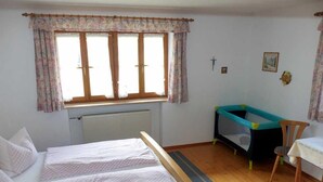 A bedroom with two beds, a window with floral curtains, a radiator, a small table, and a blue playpen. - Ferienwohnung Zuhaus, 76m², 2 Schlafzimmer, Balkon, TV, 4 Sterne, 4 Pers (Anger)