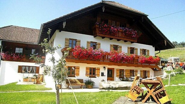 A two-story building with a balcony adorned with red flowers, a stone bench, and a wooden table in the foreground. - Ferienwohnung Zuhaus, 76m², 2 Schlafzimmer, Balkon, TV, 4 Sterne, 4 Pers (Anger)