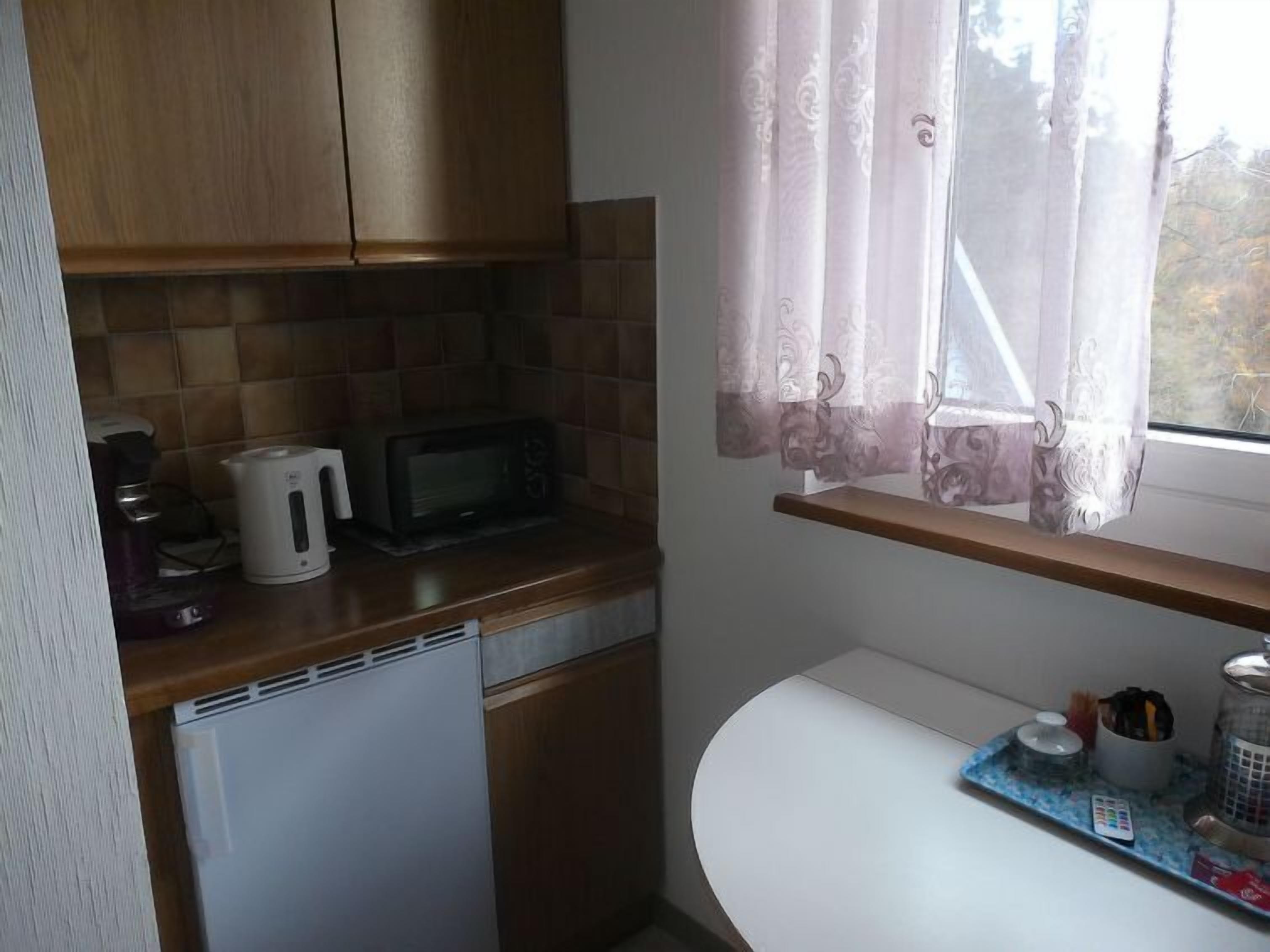 Cocina compacta con microondas, hervidor y cafetera sobre la encimera, un refrigerador pequeño y un ventanal con cortinas.