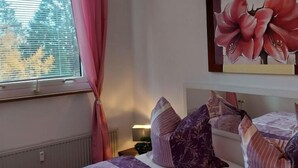 A bedroom with a bed, a window with blinds, a pink curtain, and a large floral painting on the wall. - Apartment Amina by Interhome (Braunlage)