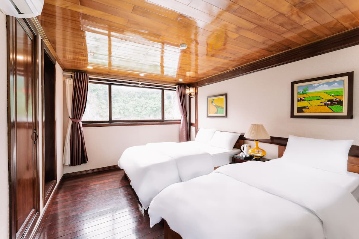 Habitación de hotel con dos camas, suelo de madera, una ventana con cortinas, un cuadro en la pared y una mesita de noche con una lámpara.