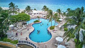 A resort pool with a view of the ocean, surrounded by palm trees and lounge chairs. - New! Modern Beach Resort - Ocean View & Access (Sunny Isles Beach)