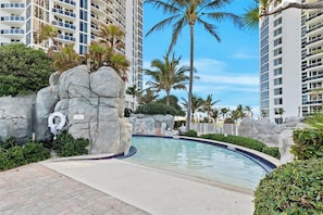 A swimming pool with a rock feature, surrounded by palm trees and a high-rise building in the background.