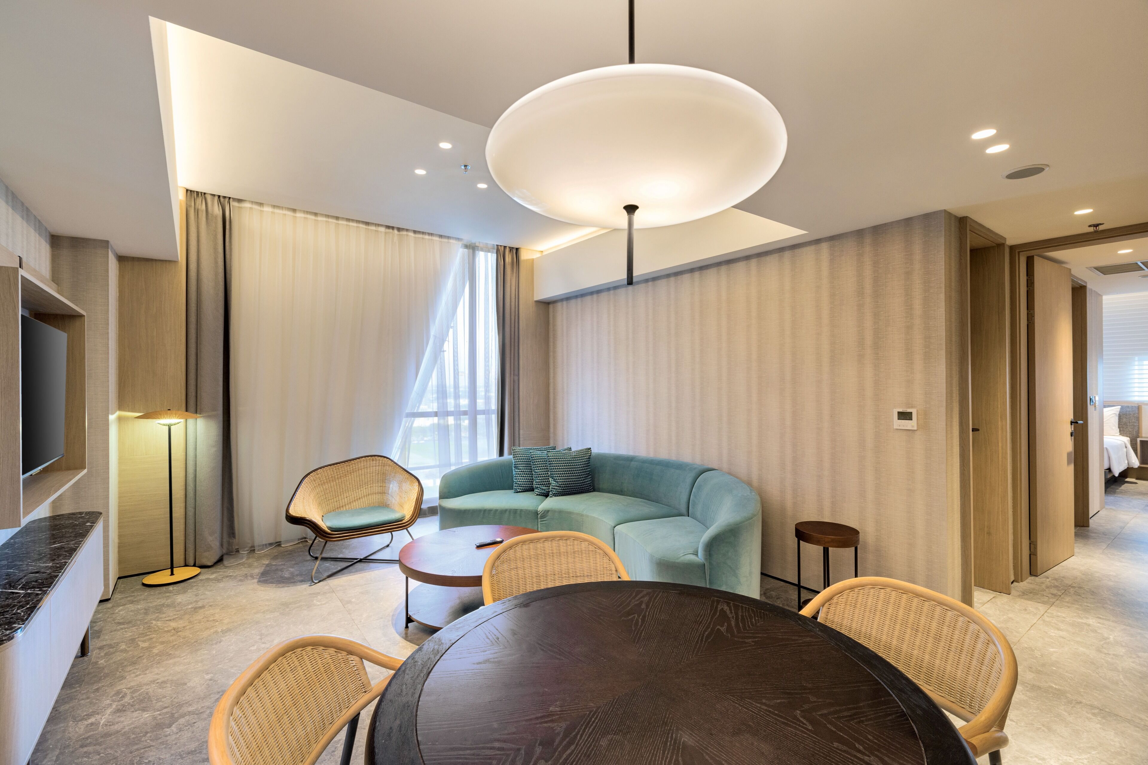 A modern hotel room with a sofa, chairs, a dining table, a TV, and a lamp.