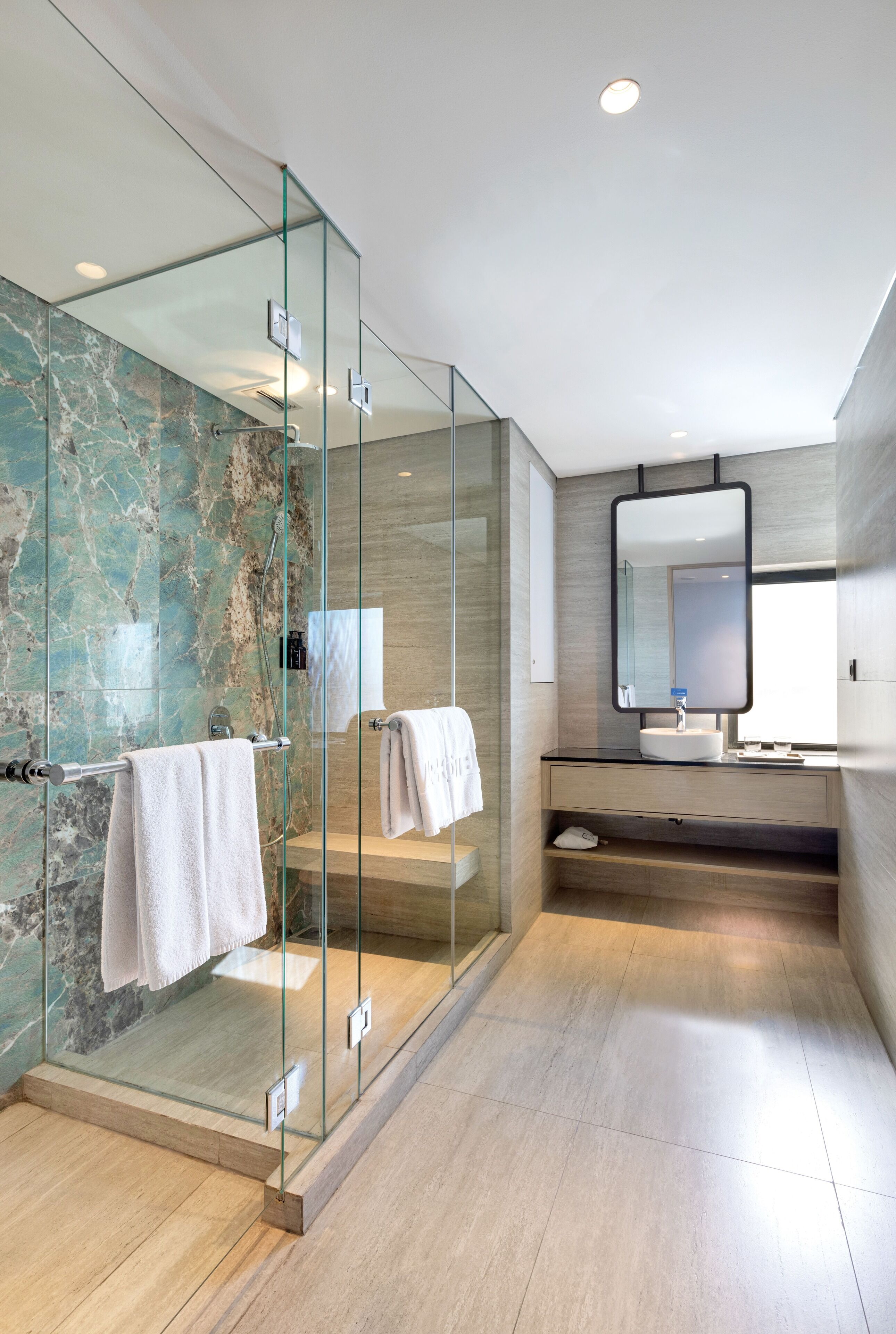 Un baño moderno con ducha de vidrio, paredes de mármol, un espejo grande y un tocador de madera.