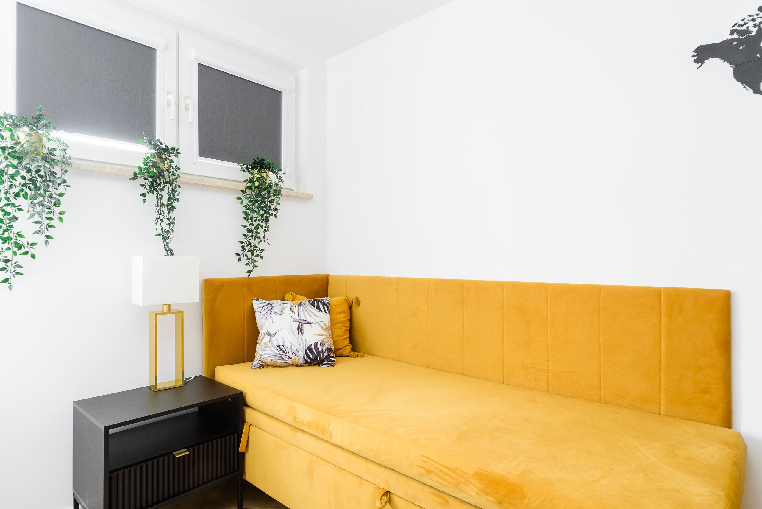 A room with a mustard-colored sofa, a black side table with a lamp, and two windows with blinds.