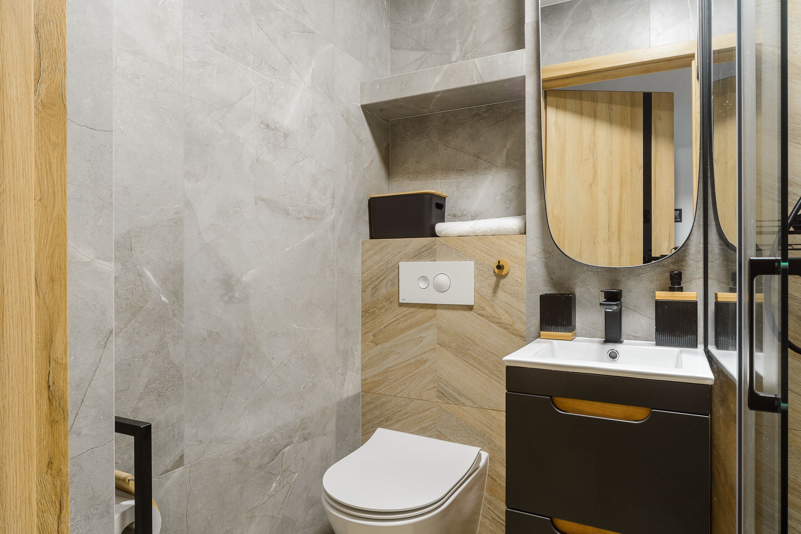 A modern bathroom with a white toilet, a wooden vanity, a mirror, and a herringbone-patterned wall.