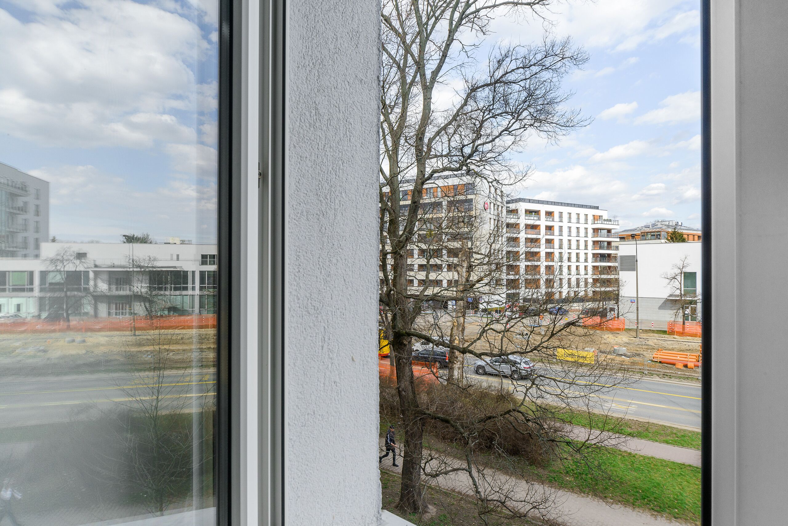 Une vue à travers la fenêtre d’un bâtiment, avec un ciel dégagé, un arbre et une route bordée de barrières de chantier.