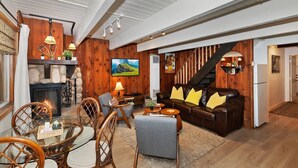 Una sala de estar acogedora con chimenea, paredes de madera, un sofá de cuero marrón con cojines amarillos, una zona de comedor con sillas de mimbre y una escalera.