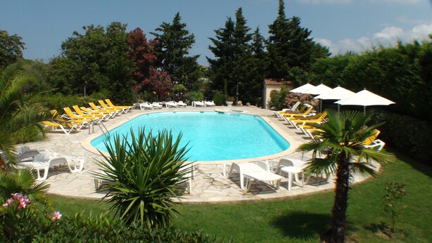 Une piscine rectangulaire entourée de chaises longues blanches et de parasols, avec une terrasse en pierre et de la verdure en arrière-plan.