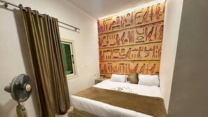 A bedroom with a bed, a standing fan, a window with curtains, and a wall with ancient Egyptian hieroglyphs. - Germania pyramids inn (Giza)