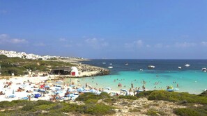 Uma praia com pessoas tomando sol e nadando, um litoral rochoso e barcos na água.