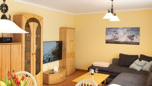 A living room with a sofa, a TV mounted on a wooden cabinet, a table with a vase of flowers, and a painting on the wall. - Apartment Ritter by Interhome (Koserow)