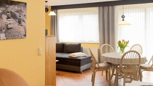 A cozy living room with a sofa, a dining table with chairs, a small kitchen area, and a large window with curtains. - Apartment Ritter by Interhome (Koserow)