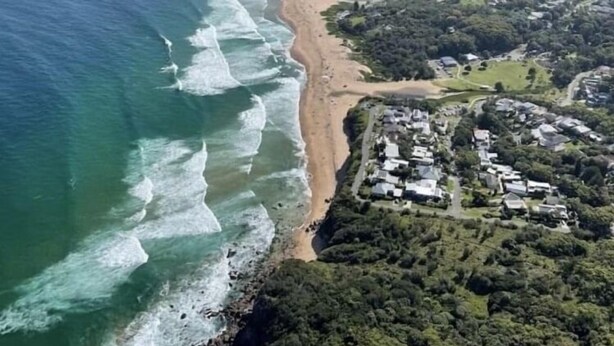 An aerial view of a coastal area with a beach, residential buildings, and surrounding hills.