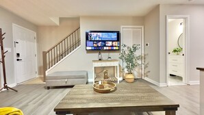 Una sala de estar con una televisión de pantalla plana montada en la pared, un banco de madera, una mesita blanca con una bandeja y una planta en maceta.