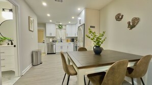 Una cocina moderna con armarios blancos, un lavavajillas de acero inoxidable y un refrigerador. Hay una mesa de comedor con sillas y un florero con flores.