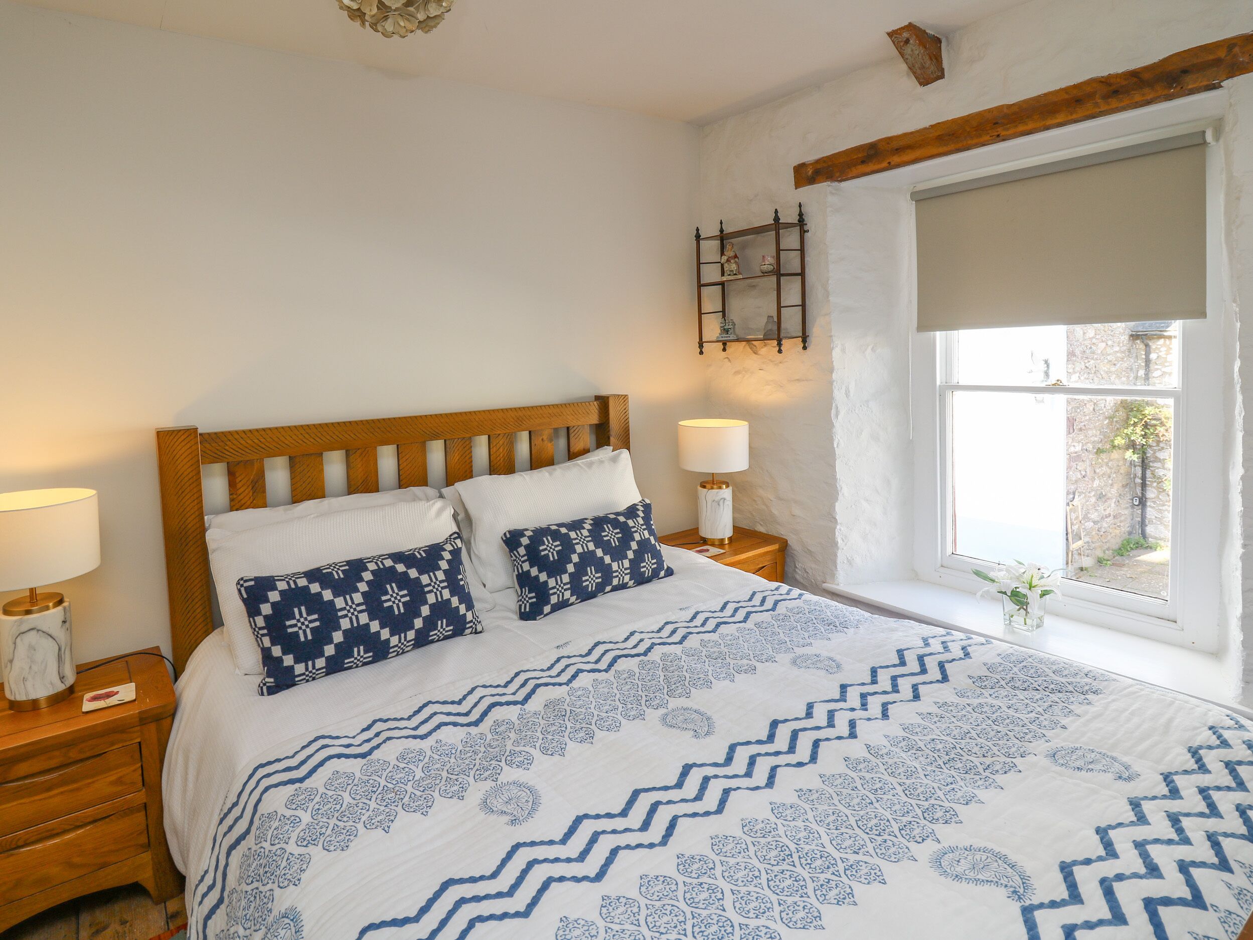 A bedroom with a wooden headboard, two bedside tables with lamps, a patterned bedspread, and a window with a view of greenery.
