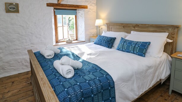 Een slaapkamer met een houten bed, witte lakens, een blauw beddegoed met patroon, en een raam met uitzicht op groen.