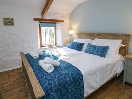 Un dormitorio con una cama de madera, sábanas blancas, una colcha de cuadros azules y un ventanal con vistas a zonas verdes.