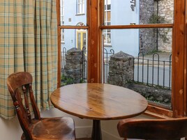 Una mesa redonda de madera con dos sillas, una cortina escocesa y vista a una calle con edificios y un muro de piedra.