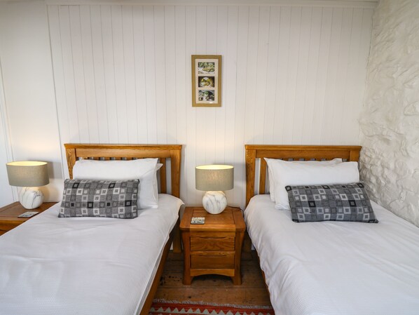 A double room with two beds, each with a wooden headboard, white bedding, and patterned pillows. There are bedside tables with lamps and a framed picture above the beds.