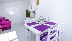 Ein heller, moderner Wohnraum mit einem weißen Sofa, einem Esstisch mit violetten Unterlegmatten und einer Küche im Hintergrund.