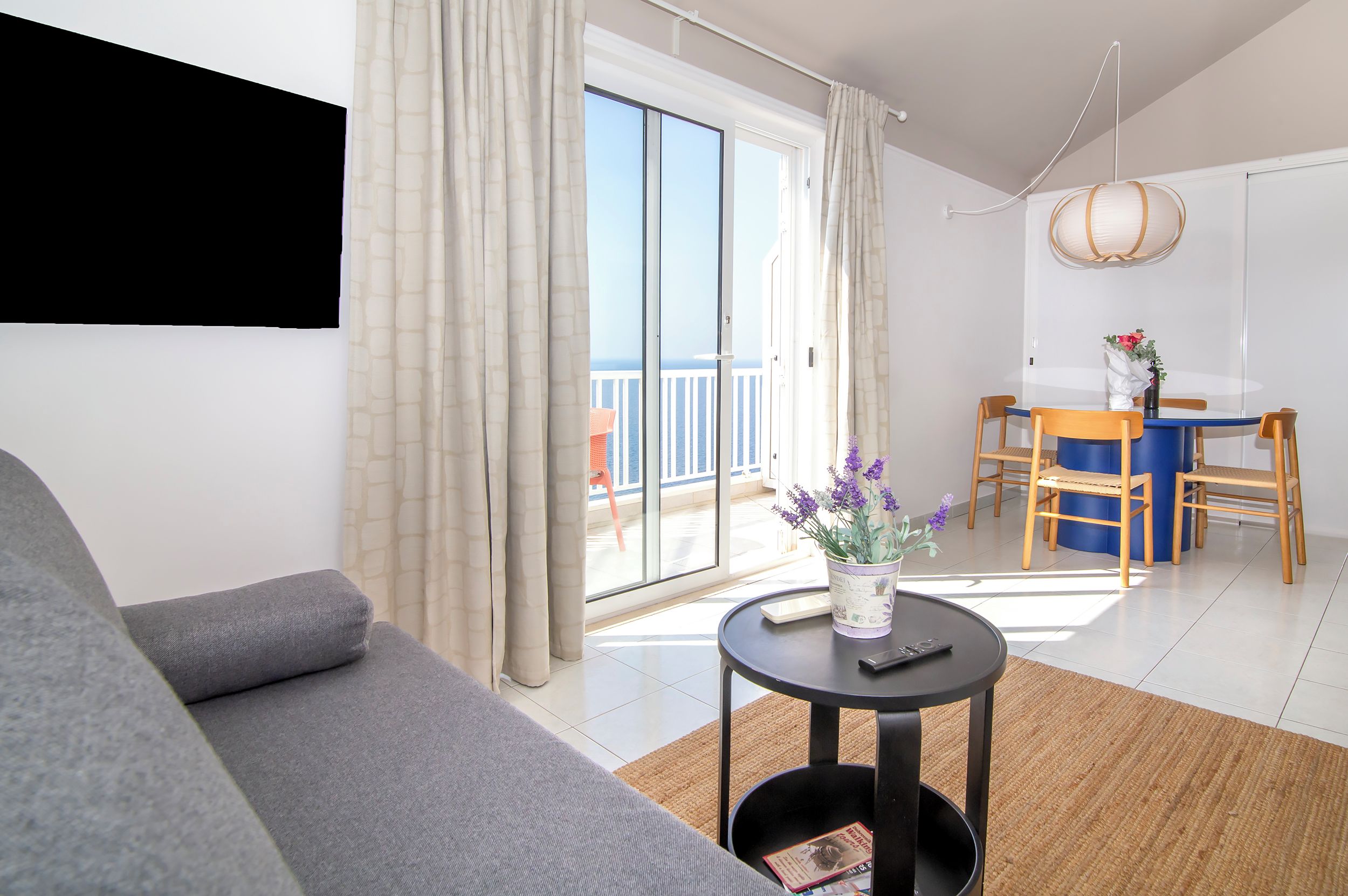 Un salon moderne avec un canapé gris, une petite table ronde ornée d’un vase de fleurs et une vue sur la mer grâce à de grandes portes coulissantes.