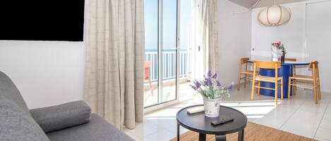 A modern living room with a grey sofa, a small round table with a vase of flowers, and a view of the sea through large sliding doors.