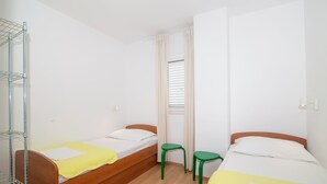 Een kleine, lichte kamer met twee éénpersoonsbedden, een raam met zonwering, een groene kruk en een metalen rek.