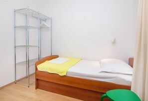 Un lit simple avec un cadre en bois, une couverture jaune et des oreillers blancs. Une étagère en métal se trouve à gauche du lit.
