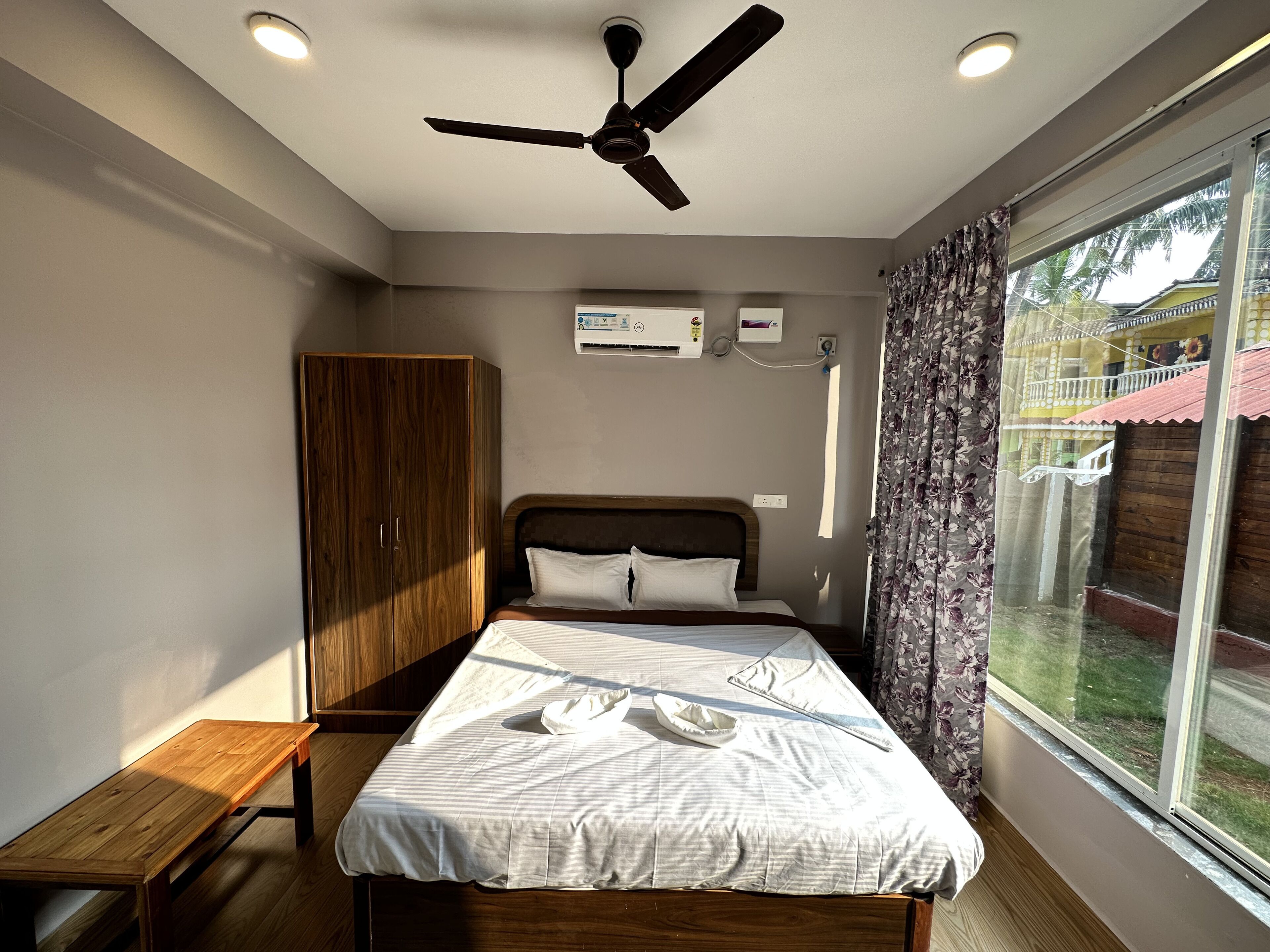 Un dormitorio con cama, armario de madera, ventilador de techo, equipo de aire acondicionado y ventana con vista a un edificio y árboles.