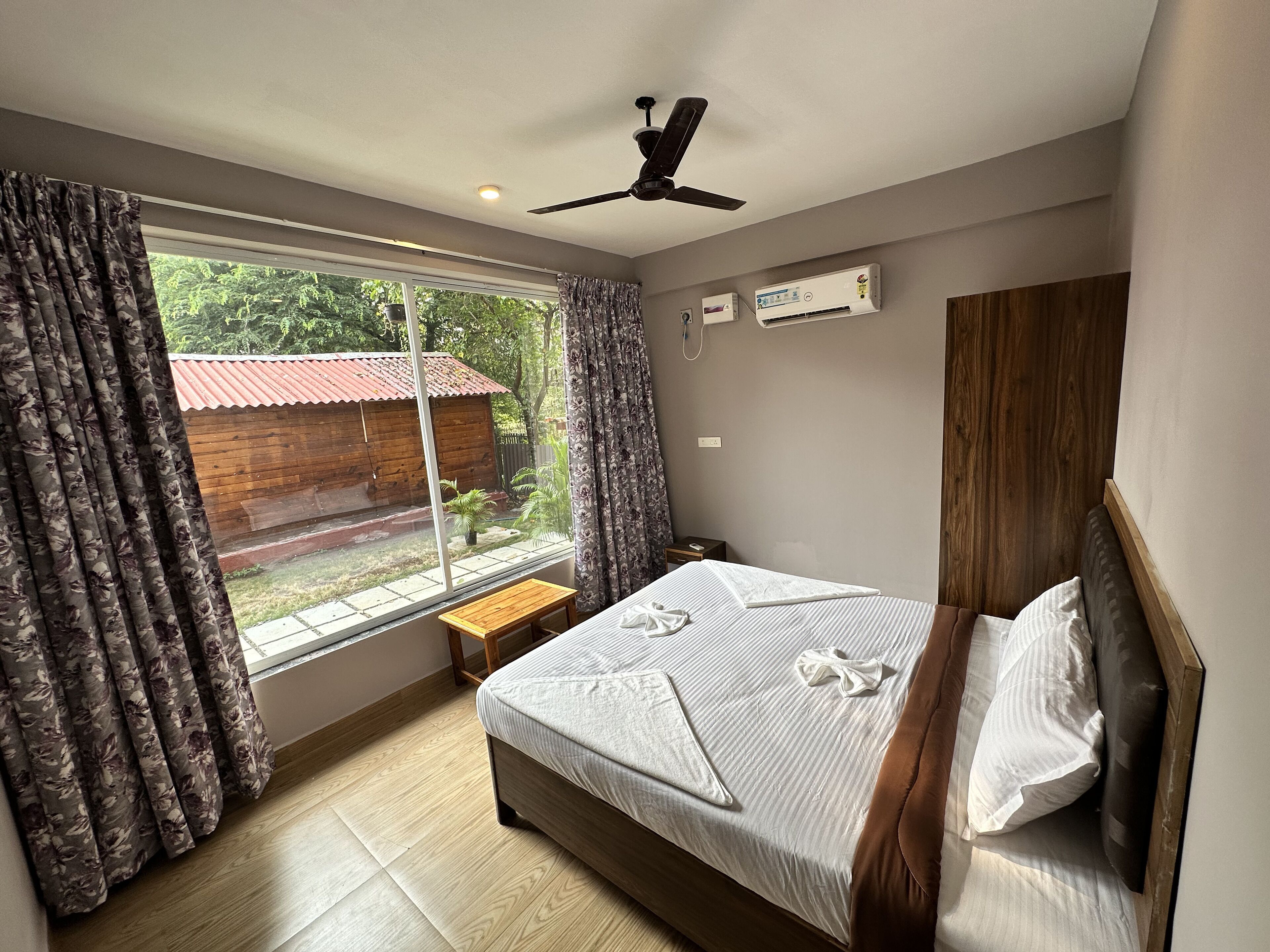 A bedroom with a bed, a ceiling fan, an air conditioning unit, a window with curtains, and a view of a wooden structure outside.