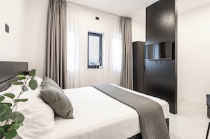 Un dormitorio moderno con una cama grande, un televisor montado en una pared oscura y una ventana con cortinas.
