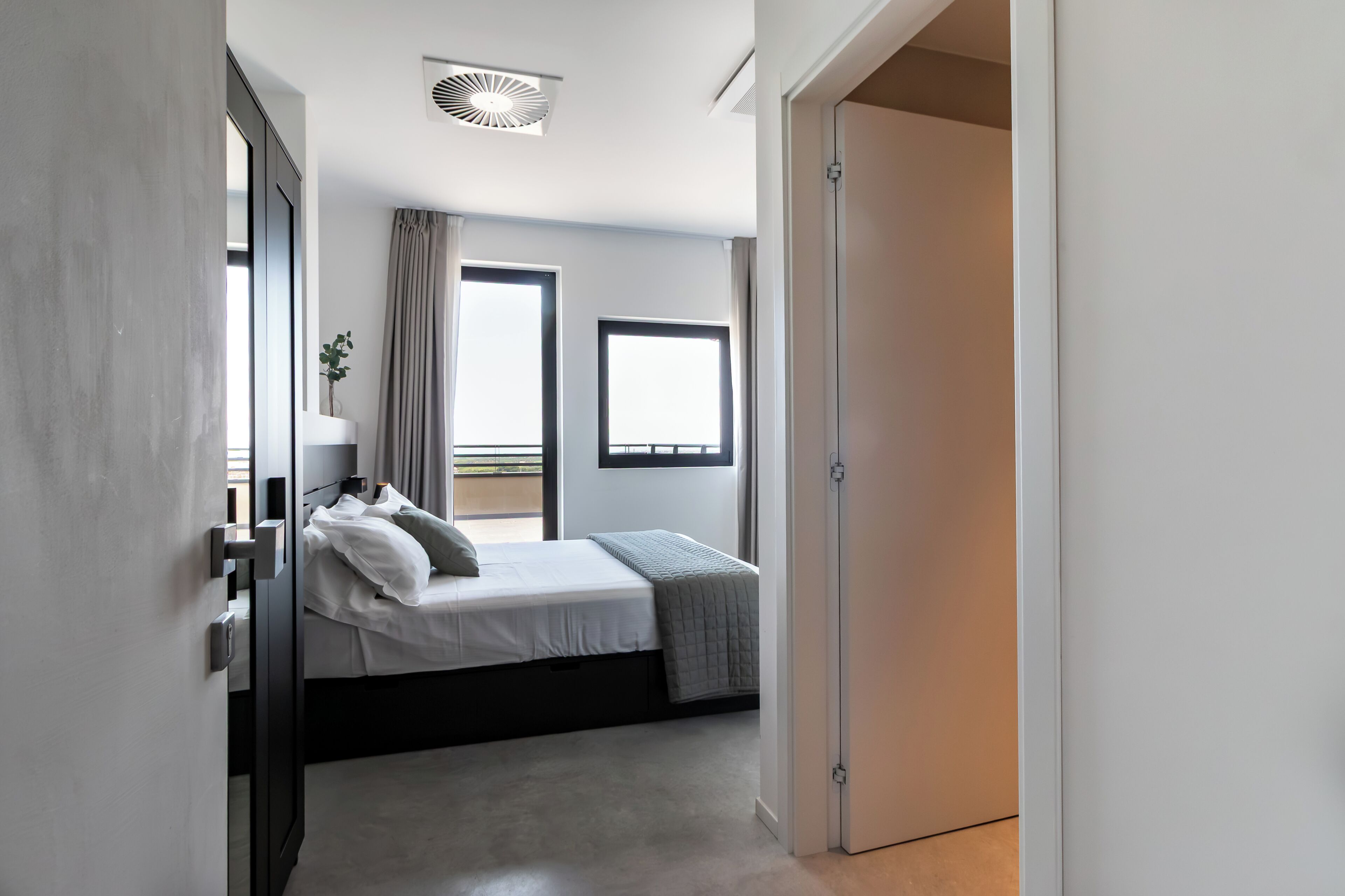 Une chambre à coucher moderne avec un lit, deux fenêtres, une plante et une porte.