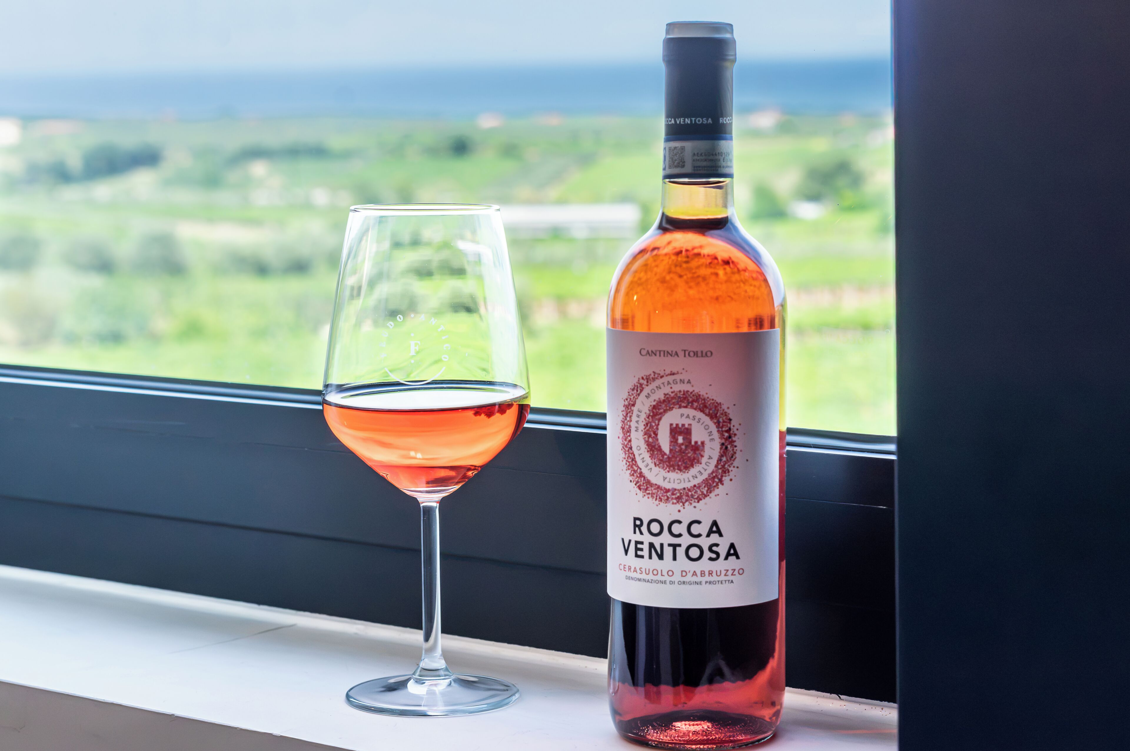 A bottle of Rocca Ventosa wine and a glass of wine on a windowsill with a view of a green landscape.