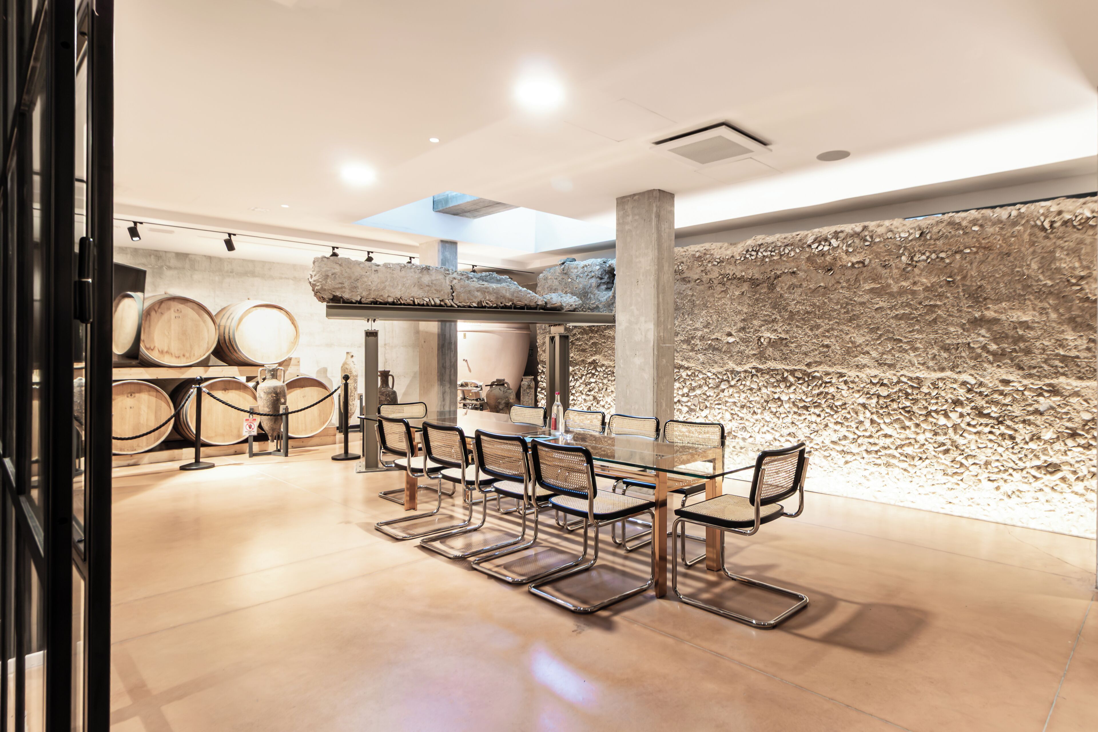 Un comedor moderno con una mesa de vidrio y sillas de metal, rodeado de barricas de vino y una pared de piedra texturizada.
