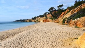 Une plage de sable fin, un ciel dégagé, une falaise rocheuse couverte de végétation et une mer calme.