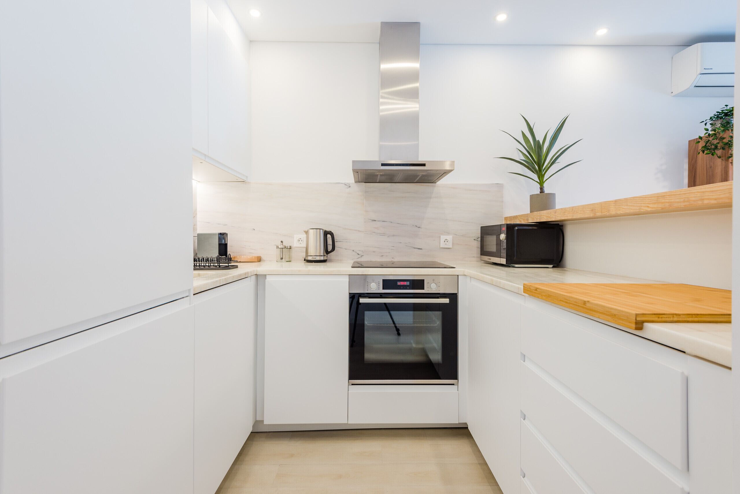 A compact kitchen with white cabinetry, a stainless steel range hood, a microwave, an oven, and a small potted plant on the countertop.