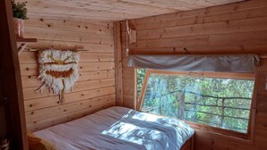 Habitación tipo cabaña de madera con cama, ventana que ofrece vistas a árboles y una planta colgante en la pared.