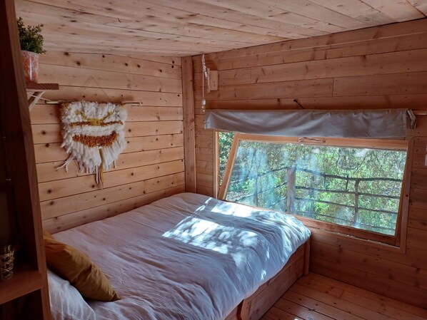 Habitación tipo cabaña de madera con cama, ventana que ofrece vistas a árboles y una planta colgante en la pared.
