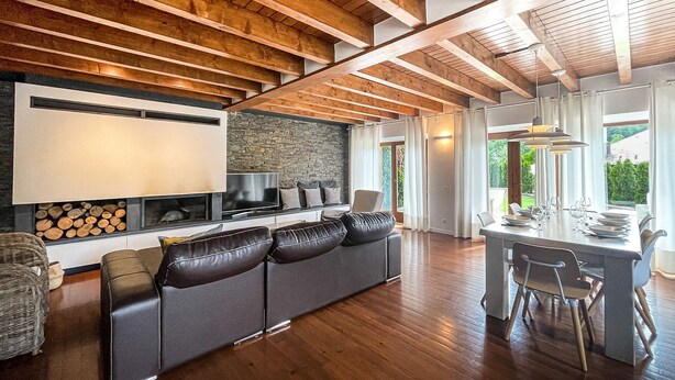 Una sala de estar moderna con una pared de piedra, vigas de madera en el techo, televisor integrado y un comedor con vistas a zonas verdes.