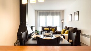 Uma área de refeições com uma mesa posta para quatro pessoas, um sofá com almofadas, uma janela com cortinas e um quadro pendurado na parede.